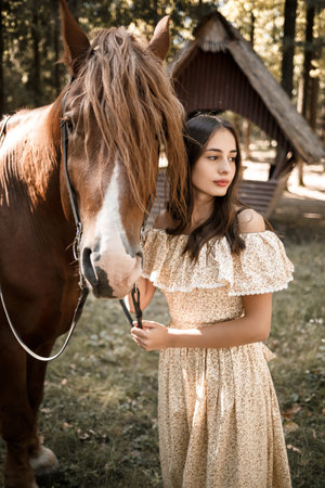 A beautiful young girl dressed in a dress stands near a horse in the forestの写真素材