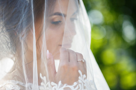 Portrait of a happy bride in a long white wedding dress and with a veil on her face in a green park on nature. Wedding image of a young girl, women's makeup and hairstyle. Marriage conceptの写真素材