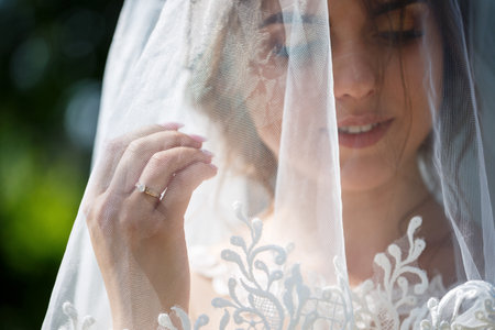 Portrait of a happy bride in a long white wedding dress and with a veil on her face in a green park on nature. Wedding image of a young girl, women's makeup and hairstyle. Marriage conceptの写真素材