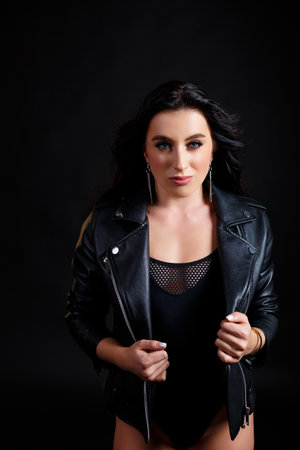 A slender tanned woman with a perfect body in a black bodysuit and a black leather biker jacket posing in the studio on a black backgroundの写真素材