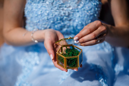 A young woman in a blue long dress sits against a green field with a casket in her hands. Fashion...