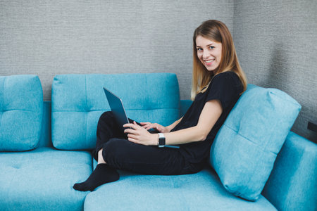 A young woman is sitting on a sofa and working on a laptop. Remote work. Work at home online