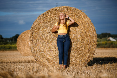 A young beautiful blonde stands on a mown wheat field near a huge sheaf of hay, enjoying nature. Nature in the villageの写真素材