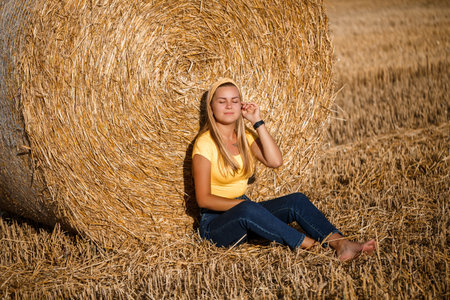 Beautiful young woman near a sheaf of hay in a field. Holidays in the village, a girl enjoying nature in a mown field on a sunny dayの写真素材