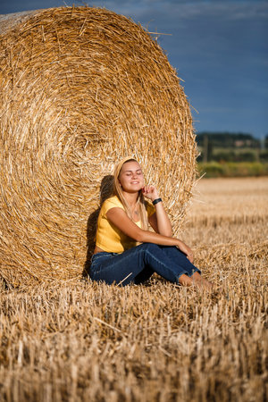 Beautiful young woman near a sheaf of hay in a field. Holidays in the village, a girl enjoying nature in a mown field on a sunny dayの写真素材