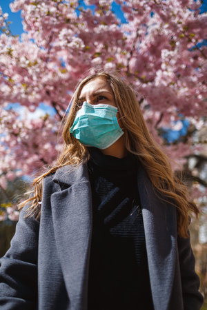 Girl, young woman in a protective sterile medical mask on her face, looking at the camera outdoors, in the spring garden. Air pollution, virus, pandemic coronavirus concept.の写真素材