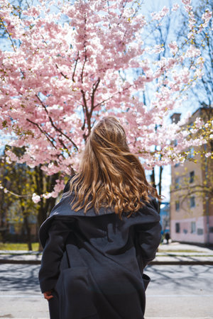 Young slender female model with long wavy hair and, dressed in a gray coat, sneakers, spinning on the street. Spring blossom trees woman girl laughs and runs to rejoiceの写真素材