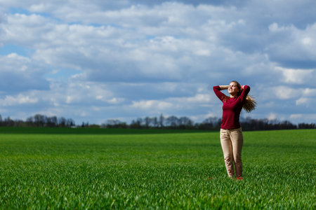 Beautiful young blonde woman stands on green grass in a park. Blue sky with clouds. The girl smiles and enjoys a good day.の写真素材
