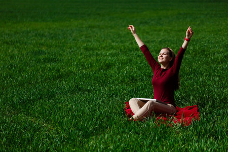 Young successful woman is sitting on green grass with a laptop in her hands. Rest after a good working day. Work on the nature. Student girl working in a secluded place. New business ideasの写真素材