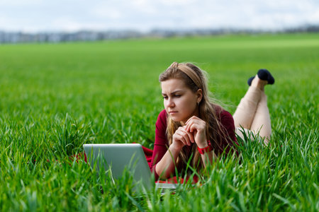 Beautiful young blonde woman is lying on the green grass in the park with a laptop and working. Blue sky with clouds. The girl smiles and enjoys a good day. Work on the nature on a sunny day.の写真素材