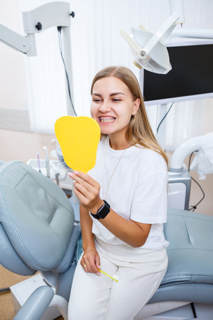 A beautiful woman in a white T-shirt sits in a dental office, looks in the mirror, smiles and holds a toothbrush in her hands. Girl with braces shows oral care. Dentistry, dental treatmentの写真素材