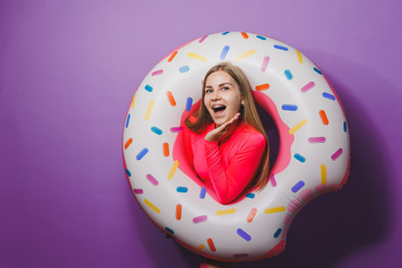 Cheerful slender woman with long hair holding an inflatable ring on a purple background. Attractive girl in a pink swimsuitの写真素材