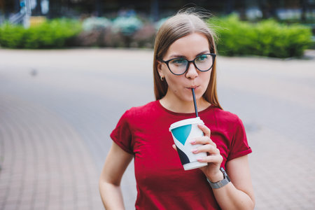 Happy young female with backpack smiling and looking away while walking on city street and enjoying fresh coffee to goの写真素材