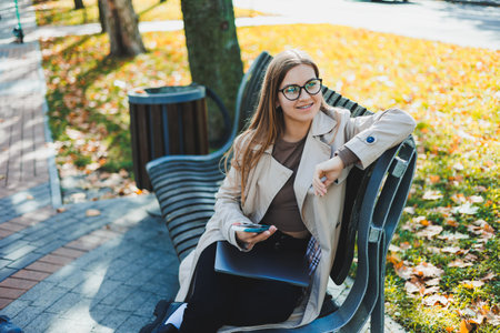 A smiling woman works with a laptop on a bench in the Autumn Park. She has a great smile, long hair and big blue eyes. Portrait of a modern working woman. Yellow park backgroundの写真素材
