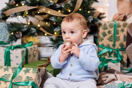 A one-year-old boy sits near a decorated Christmas tree with gifts. Christmas tree in the house. A...