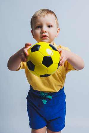 A little boy in a yellow t-shirt with a soccer ball in his hand smiles isolated on a white background. Sports child holding a ball. Children's sports game. Little athlete.の写真素材
