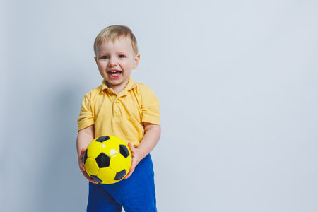 A little boy with a soccer ball in his hands on a white background, a child is a novice football player, a sport for children. Little athlete. Yellow and blue football kit for kidsの写真素材