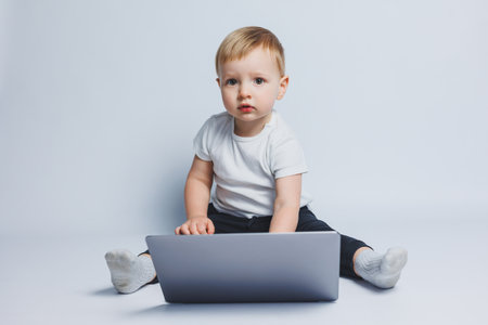 A smart boy 3-4 years old sits with a laptop on a white background. A child in a white T-shirt and...