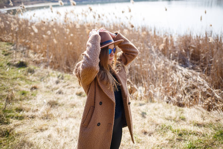 A girl in a brown coat, hat and glasses walks in a park with a lake under the bright sun. Rejoices in life and smiles. The beginning of springの写真素材