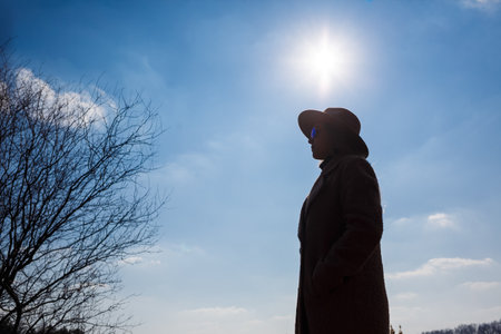 Silhouette of a girl in a hat and coat on a background of the sky with clouds and the sun.の写真素材