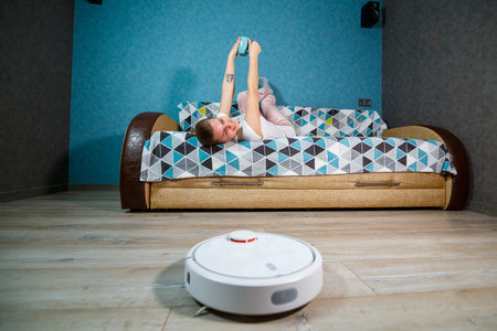 Simple and easy cleaning with modern technology for the household. Girl drinks tea and rests while the vacuum cleaner does the cleaning of the houseの写真素材