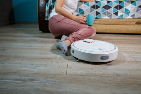 Simple and easy cleaning with modern technology for the household. Girl drinks tea and rests while the vacuum cleaner does the cleaning of the houseの写真素材