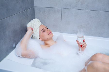 Young beautiful woman with a white towel on her head lies in a white bath. In the hands of a glass...