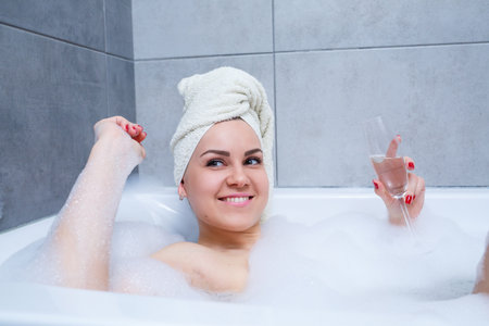 Girl woman with a white towel on her head lies with a glass of champagne in a white bathtub. In her...
