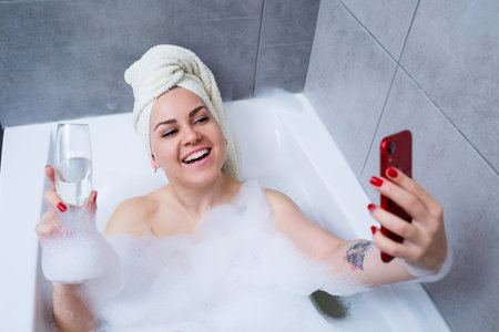 Girl woman with a towel on her head lies with a glass of champagne in a white bathtub and takes a...