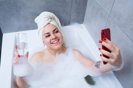A beautiful woman with a towel on her head lies with a glass of champagne in a white bath and takes...