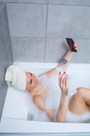 Girl woman with a towel on her head lies with a glass of champagne in a white bathtub and takes a...