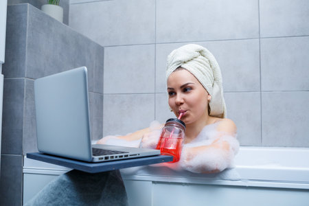 A young woman with a towel on her head drinks juice watching a movie on a laptop while sitting in a...