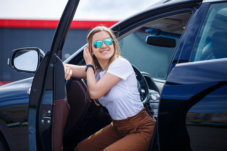 Young beautiful woman with long hair sits in a black car at a parking lot. Pretty girl in casual...