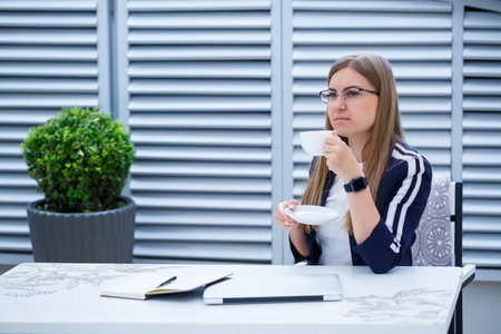 Beautiful young woman in a white T-shirt is drinking coffee and working on a laptop and smiling...