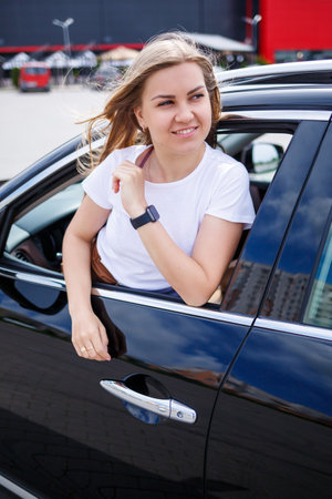 Young beautiful woman with long hair sits in a black car at a parking lot. Pretty girl in casual...