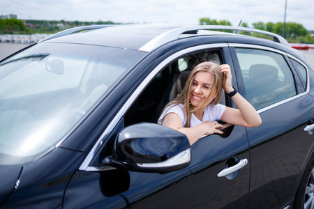 Young beautiful woman with long hair sits in a black car at a parking lot. Pretty girl in casual...