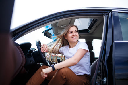 Young beautiful woman with long hair sits in a black car at a parking lot. Pretty girl in casual...