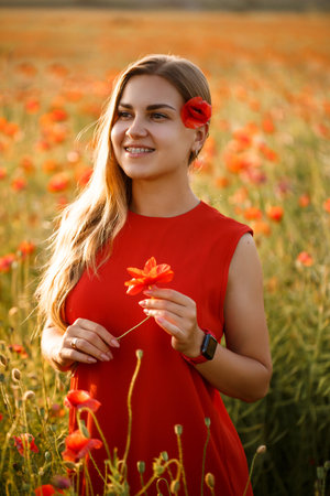 Happy blonde woman in red dress holding a flower in her hair on a poppy field. Fun summer vibes. Relaxed atmosphere, positive emotions.の写真素材