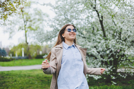 Back view girl walking under blossoming apple tree in natural park on hill slope in Prague. Elegant young lady in hat and brown coat enjoying flowering garden at sunny spring day.の写真素材