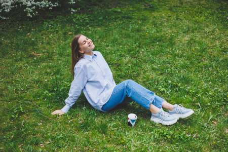 Young hipster woman in denim jacket is resting on green lawn in park, drinking coffee, relaxing...