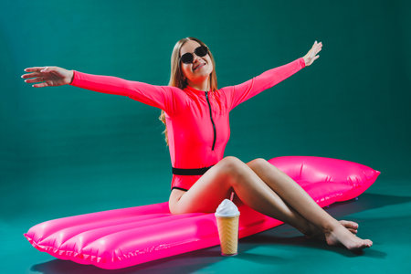 Pretty slim woman in a pink swimsuit lies on an inflatable mattress with a cocktail in her hands, on an isolated green background. Summer vacation vacation at sea tanning concept. Pink air mattressの写真素材