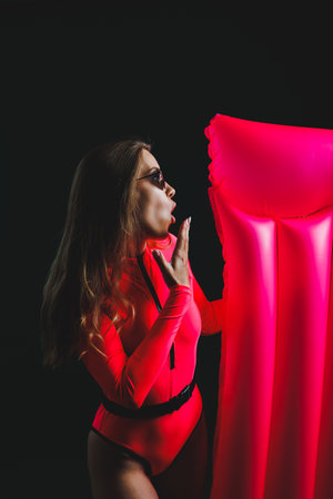Young woman in a pink swimsuit, isolated on a dark background. Vacation lifestyle concept, summer holidays. A girl in sunglasses with joyful emotion holds an inflatable pink mattress in her handsの写真素材