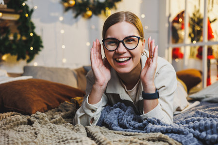 Happy smiling woman in glasses lies on the bed on the background of Christmas decorations. The woman is happy with the Christmas holidays. New Year's atmosphere at home.の写真素材
