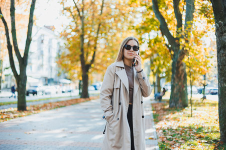 Young smiling woman walking in autumn park, talking on smartphone, wearing trench coat, sunglasses, happy mood, fashion style trendの写真素材