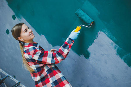 Young smiling pretty woman in shirt holding roller in right hand and painting wall. Repair in the room.の写真素材