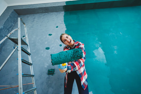 An emotional woman paints the walls. Young smiling pretty woman in shirt holding roller in right hand and singing. Repair in the room.の写真素材