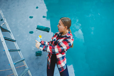 An emotional woman paints the walls. Young smiling pretty woman in shirt holding roller in right hand and singing. Repair in the room.の写真素材