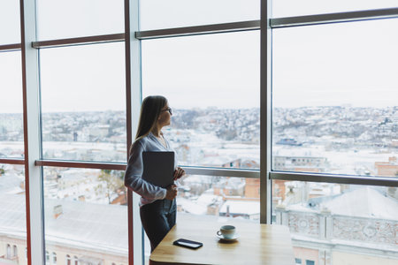 Cheerful young fair-haired business woman of European appearance in a white shirt and glasses holds a portable laptop in her hands, stands near a large window in the office on the top floorの写真素材