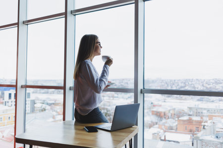 European business woman using a laptop computer while working. The concept of a modern successful woman. Young beautiful smiling woman with coffee sitting in a cafe. Open office interiorの写真素材