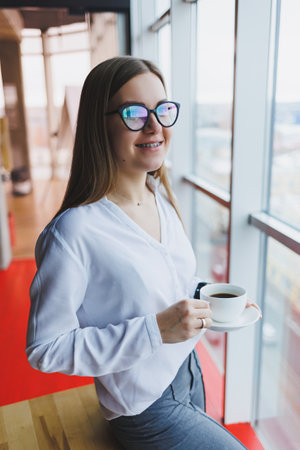 A young woman in glasses of European appearance, she is in a white shirt drinking coffee and looking...